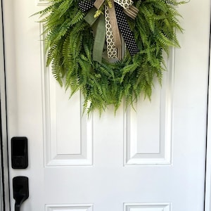 May include: A circular green fern wreath featuring a decorative bow made of multiple ribbons in tan, olive green, black with white polka dots, and a white geometric pattern, hanging on a white door with a black handle and electronic lock.