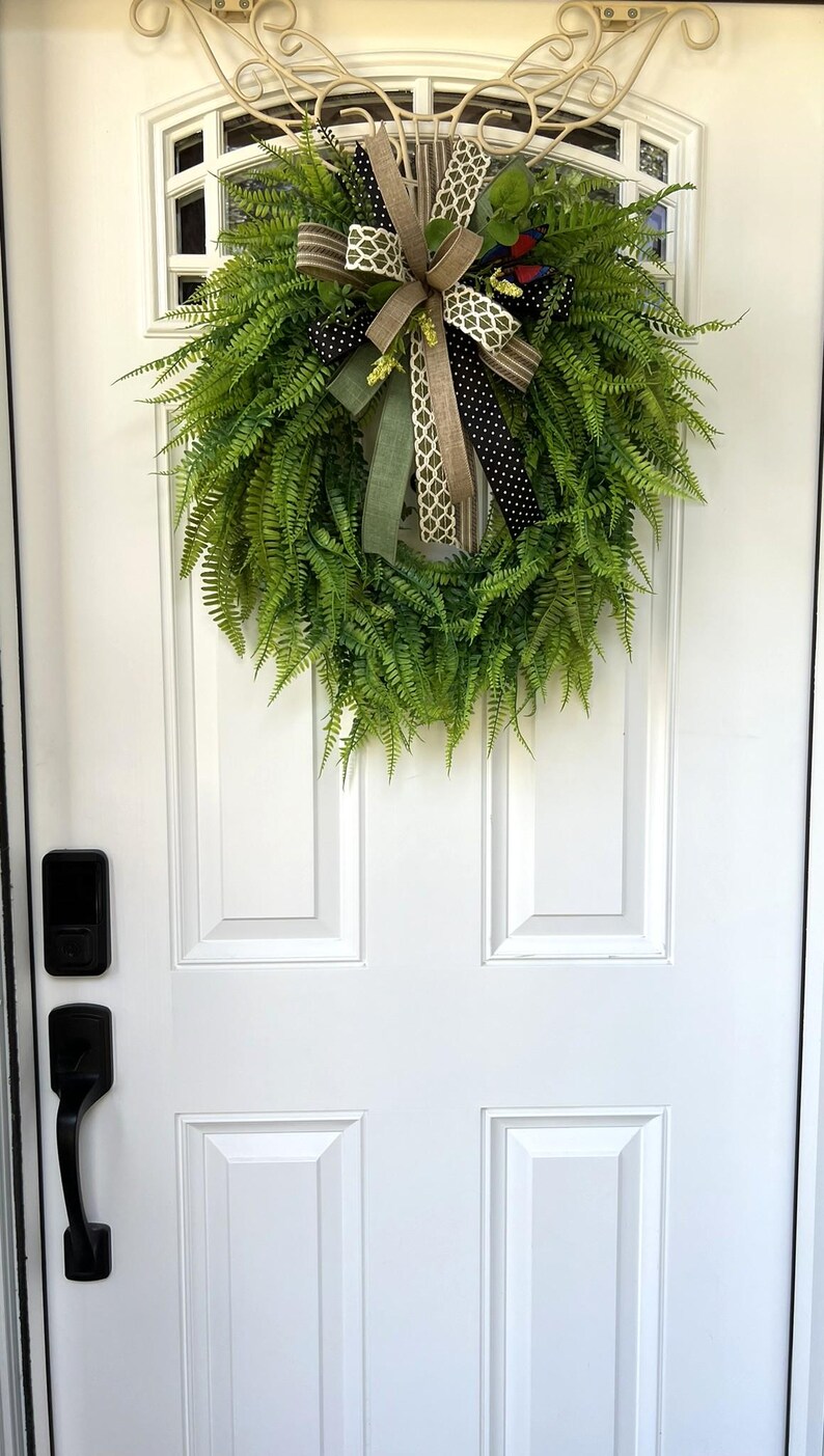 May include: A circular green fern wreath featuring a decorative bow made of multiple ribbons in tan, olive green, black with white polka dots, and a white geometric pattern, hanging on a white door with a black handle and electronic lock.