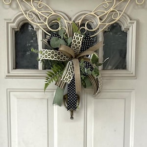 May include: A decorative door swag featuring a mix of green fern and eucalyptus foliage, accented with multiple ribbons in tan, olive green, and black with white polka dots, all suspended from a cream-colored metal scrollwork door hanger.