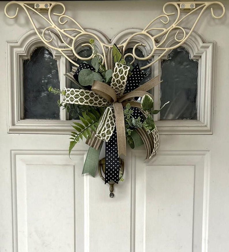 May include: A decorative door swag featuring a mix of green fern and eucalyptus foliage, accented with multiple ribbons in tan, olive green, and black with white polka dots, all suspended from a cream-colored metal scrollwork door hanger.