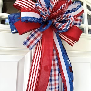Op de afbeelding: Een decoratieve strik in een patriottisch kleurenschema van rood, wit en blauw. De strik is gemaakt van meerdere linten met diverse patronen, waaronder effen rood, rood-witte strepen, blauw met glinsterende sterren en een rood-wit-blauw ruitpatroon.