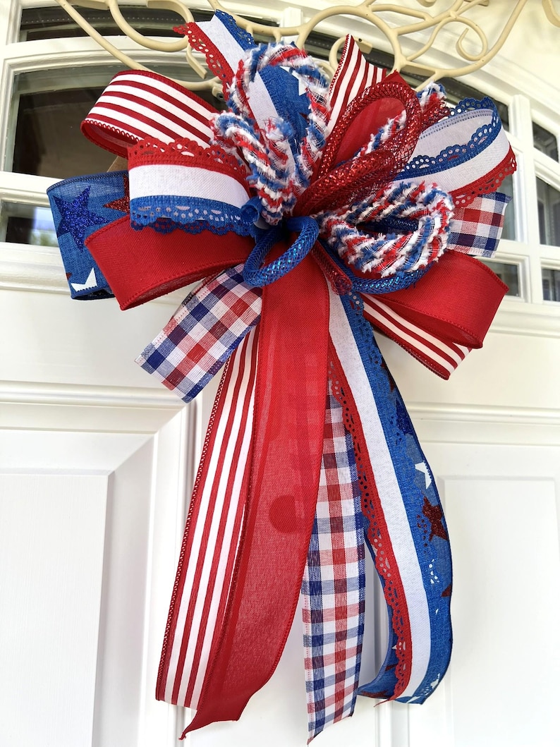 Op de afbeelding: Een decoratieve strik in een patriottisch kleurenschema van rood, wit en blauw. De strik is gemaakt van meerdere linten met diverse patronen, waaronder effen rood, rood-witte strepen, blauw met glinsterende sterren en een rood-wit-blauw ruitpatroon.