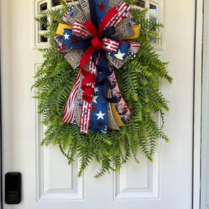 May include: A patriotic wreath with a fern base and a large bow. The bow features red, white, and blue ribbons with stars, stripes, and text. The wreath is hanging on a white door.