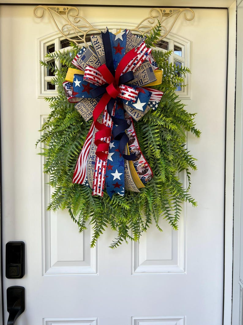 May include: A patriotic wreath with a fern base and a large bow. The bow features red, white, and blue ribbons with stars, stripes, and text. The wreath is hanging on a white door.