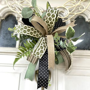 May include: A decorative bow featuring a mix of ribbons in solid tan, black with white polka dots, and olive green with a white lattice pattern. The bow is accented with artificial greenery, including fern fronds and eucalyptus leaves.