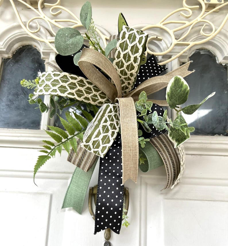May include: A decorative bow featuring a mix of ribbons in solid tan, black with white polka dots, and olive green with a white lattice pattern. The bow is accented with artificial greenery, including fern fronds and eucalyptus leaves.