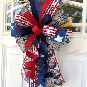 May include: A patriotic bow featuring red, white, and blue ribbons with star and stripe patterns. The bow includes ribbons with eagle illustrations and text. The bow is likely for a door or wreath decoration.