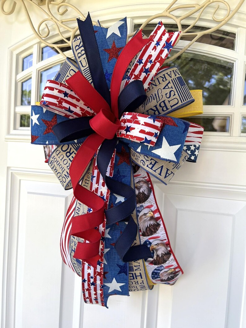 May include: A patriotic bow featuring red, white, and blue ribbons with star and stripe patterns. The bow includes ribbons with eagle illustrations and text. The bow is likely for a door or wreath decoration.