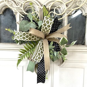 May include: A decorative door swag featuring a mix of green fern fronds, eucalyptus leaves, and small yellow floral sprigs. The arrangement is accented with multiple ribbons, including black with white polka dots, solid tan, and a white lattice pattern.