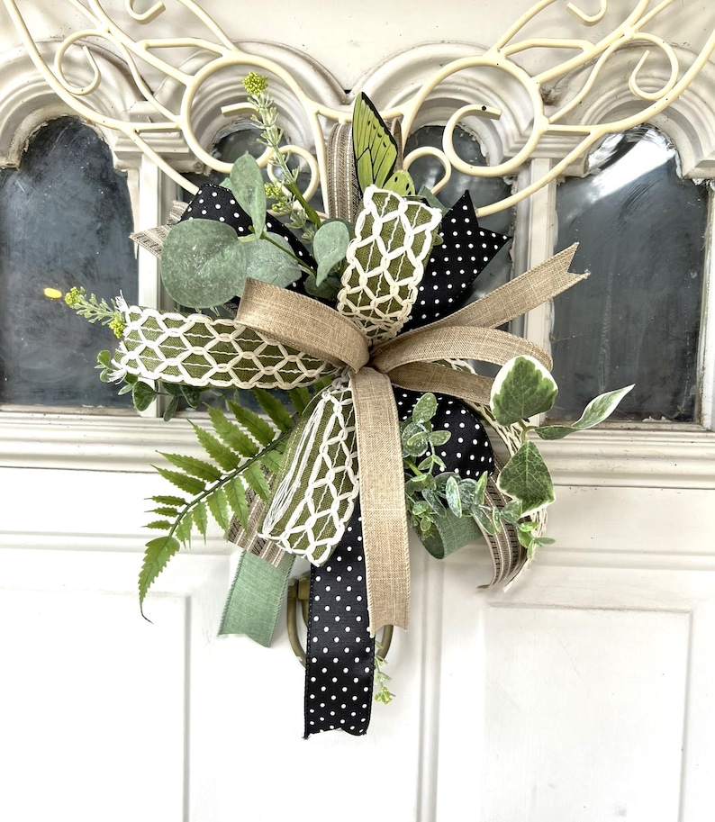 May include: A decorative door swag featuring a mix of green fern fronds, eucalyptus leaves, and small yellow floral sprigs. The arrangement is accented with multiple ribbons, including black with white polka dots, solid tan, and a white lattice pattern.