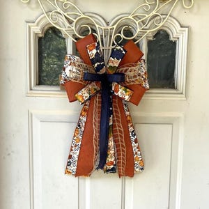 May include: A decorative bow made from multiple ribbons featuring solid burnt orange, navy blue, and a patterned ribbon with autumn leaves and pumpkins. The bow includes textured burlap accents and long, flowing tails hanging downwards.
