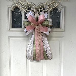 May include: A pink, green, and white bow with a burlap ribbon is attached to a white metal angel door hanger.