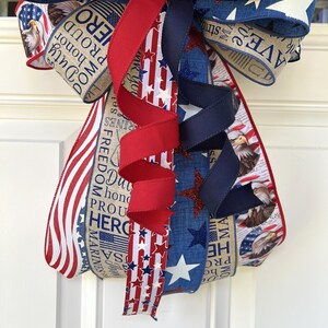 May include: A patriotic bow with red, white, and blue ribbons. The bow features star and stripe patterns, along with text that reads "Duty, Honor, Proud, Hero." The bow is likely for a wreath or other decorative use.