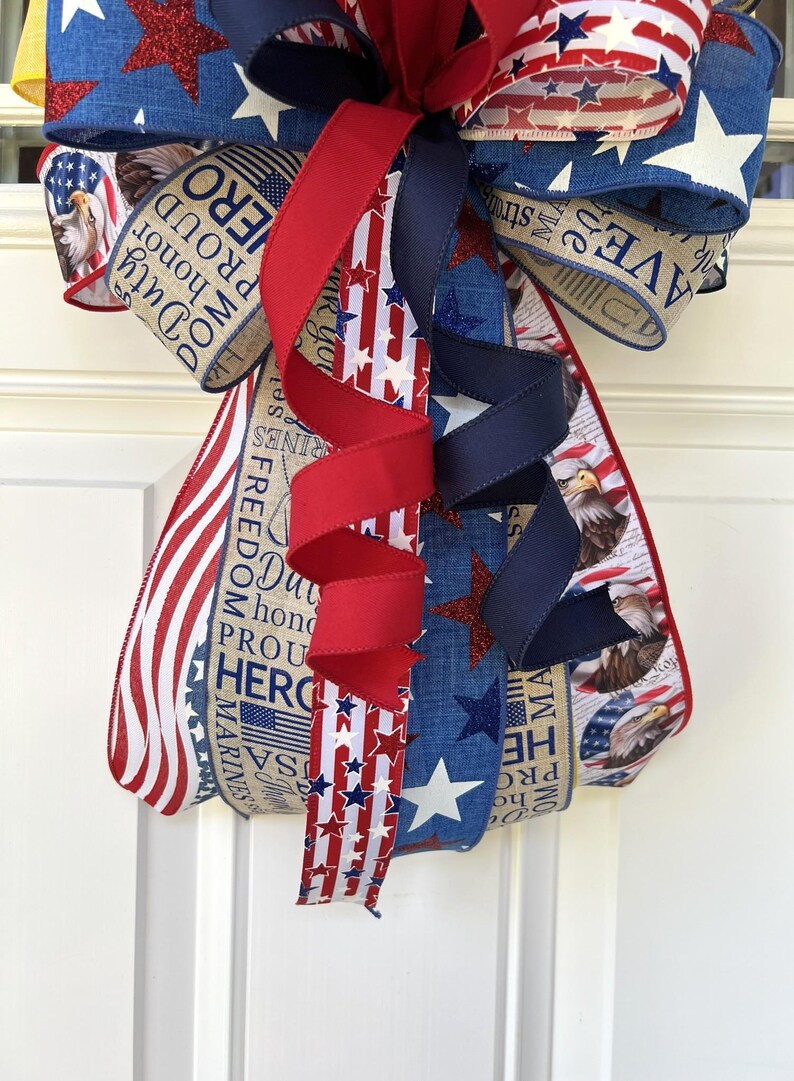 May include: A patriotic bow with red, white, and blue ribbons. The bow features star and stripe patterns, along with text that reads "Duty, Honor, Proud, Hero." The bow is likely for a wreath or other decorative use.