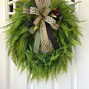 May include: A lush green fern wreath featuring a decorative multi-layered bow. The bow consists of ribbons in solid tan, olive green, black with white polka dots, and a cream-colored geometric lattice pattern, accented by small red and blue butterflies.