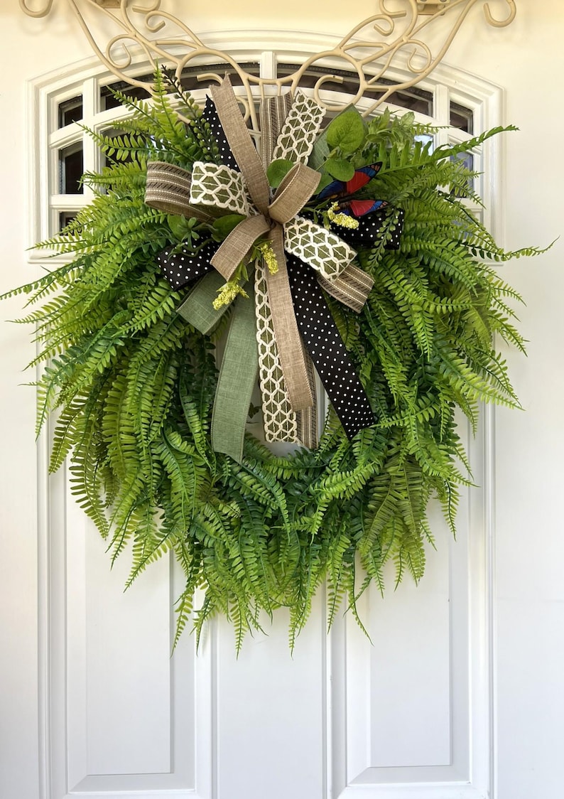 May include: A lush green fern wreath featuring a decorative multi-layered bow. The bow consists of ribbons in solid tan, olive green, black with white polka dots, and a cream-colored geometric lattice pattern, accented by small red and blue butterflies.