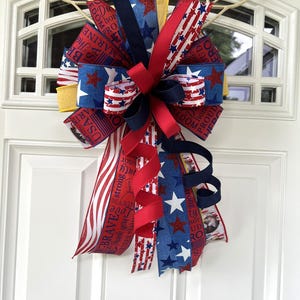 May include: A patriotic door bow featuring red, white, and blue ribbons with star and stripe patterns. Some ribbons have text that reads "USA", "Brave", "Marine", and "Proud". The bow is attached to a white door.