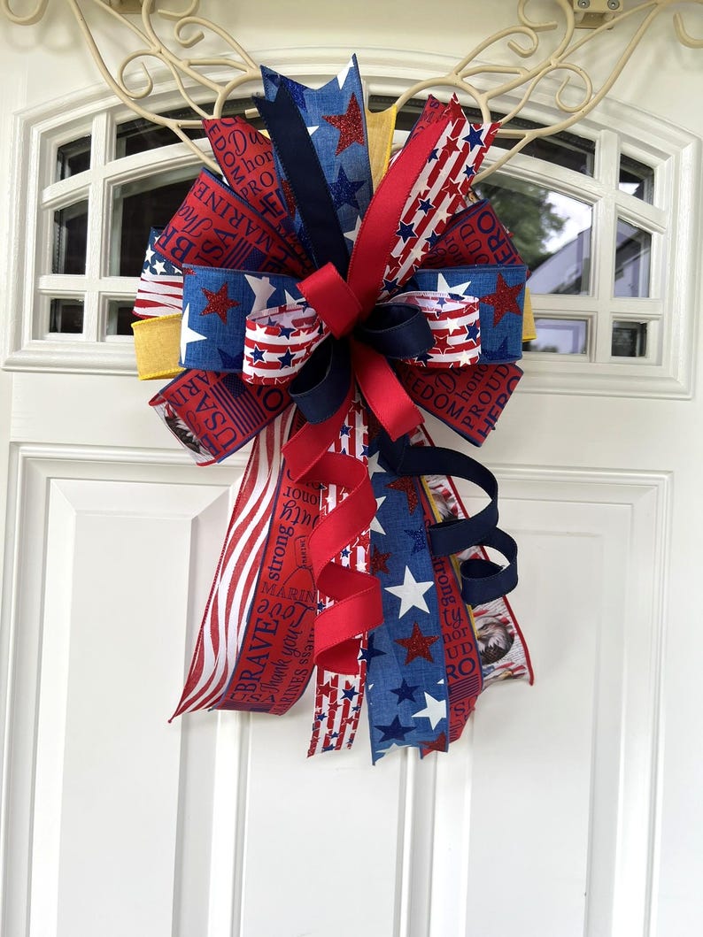 May include: A patriotic door bow featuring red, white, and blue ribbons with star and stripe patterns. Some ribbons have text that reads "USA", "Brave", "Marine", and "Proud". The bow is attached to a white door.
