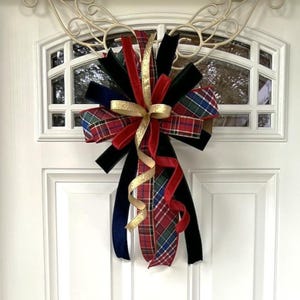 May include: A decorative bow made of multiple ribbons in solid black, solid red, and a multi-colored plaid pattern featuring red, green, blue, and white. The bow includes long, trailing ribbon tails and a central accent of shimmering gold ribbon.