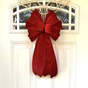 May include: A large, plush, red velvet bow hangs on a white door. The bow is attached to a decorative, ornate, cream-colored metal wreath. The bow's long, cascading tails add a touch of elegance to the door's design.