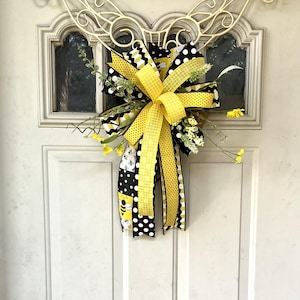 May include: A yellow and black bee-themed bow with a yellow and white checkered ribbon. The bow is attached to a white door with a decorative metal hook.