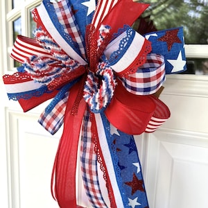 Op de afbeelding: Een decoratieve strik gemaakt van meerdere linten in rood, wit en blauw. De linten hebben diverse patronen, waaronder effen kleuren, rood-witte strepen, blauw-witte vichy-ruitjes en blauwe stof met witte en rode glittersterren.