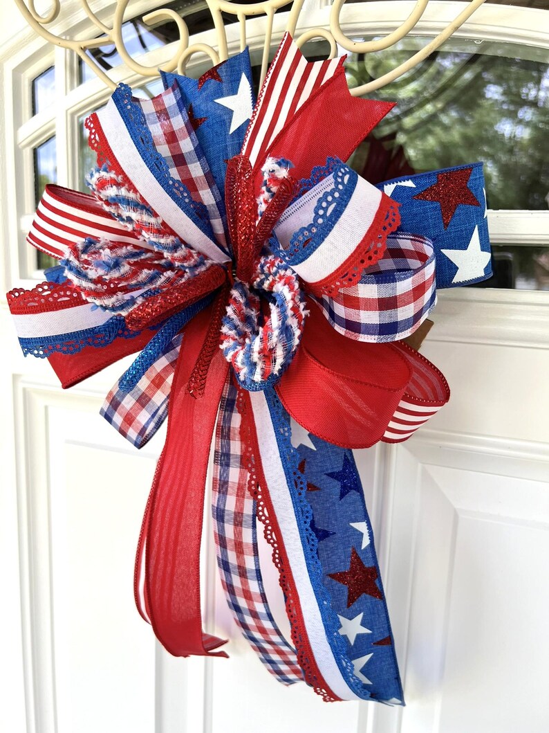 Op de afbeelding: Een decoratieve strik gemaakt van meerdere linten in rood, wit en blauw. De linten hebben diverse patronen, waaronder effen kleuren, rood-witte strepen, blauw-witte vichy-ruitjes en blauwe stof met witte en rode glittersterren.