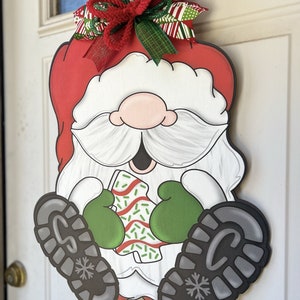 May include: A Christmas door hanger featuring a cartoon Santa Claus wearing a red hat, white beard, and green mittens. He is holding a candy cane with green sprinkles. The hanger is decorated with a red, green, and white bow.