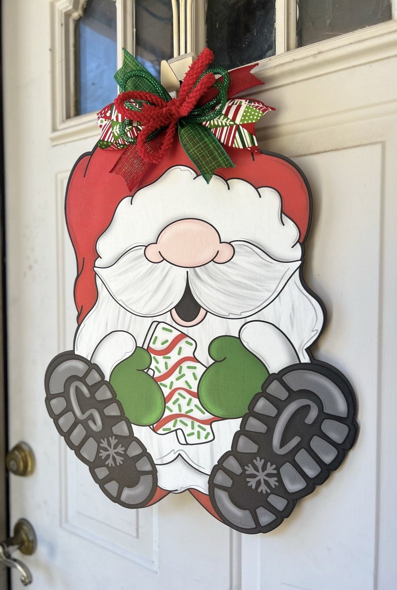 May include: A Christmas door hanger featuring a cartoon Santa Claus wearing a red hat, white beard, and green mittens. He is holding a candy cane with green sprinkles. The hanger is decorated with a red, green, and white bow.