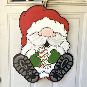 May include: A red and white Santa Claus door hanger with a green hat and a white beard. He is holding a green and white candy cane in his hands.