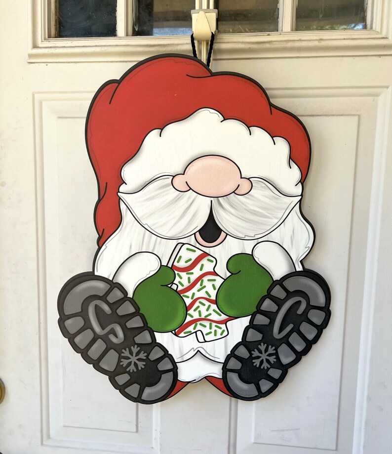 May include: A red and white Santa Claus door hanger with a green hat and a white beard. He is holding a green and white candy cane in his hands.