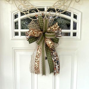 May include: A decorative door bow featuring a large multi-layered bow with various ribbons. The ribbons include patterns of leaves, plaid, and a print with figures. Colors include green, brown, and cream. The bow is attached to a white door with a decorative metal accent.