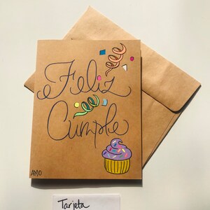 Pu&ograve; includere: Un biglietto marrone con le parole "Feliz Cumple" scritte in corsivo nero. Il biglietto &egrave; decorato con coriandoli colorati e un cupcake viola con codette. Il biglietto &egrave; etichettato "Tarjeta Sobre (en blanco)"