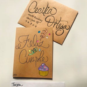Pu&ograve; includere: Un biglietto marrone con le parole "Feliz Cumple" scritte in corsivo nero. Il biglietto &egrave; decorato con coriandoli colorati e un cupcake viola con codette rosa e gialle. Il biglietto &egrave; indirizzato a "Cecilia Ortega" con l'indirizzo "45678 Broadway, New York, NY 7-8-9-1-0" scritto su una busta marrone.