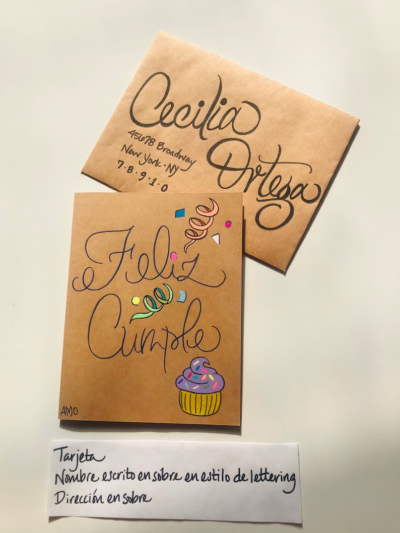 Pu&ograve; includere: Un biglietto marrone con le parole "Feliz Cumple" scritte in corsivo nero. Il biglietto &egrave; decorato con coriandoli colorati e un cupcake viola con codette rosa e gialle. Il biglietto &egrave; indirizzato a "Cecilia Ortega" con l'indirizzo "45678 Broadway, New York, NY 7-8-9-1-0" scritto su una busta marrone.