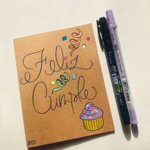Pu&ograve; includere: Un biglietto marrone con le parole "Feliz Cumple" scritte in inchiostro nero. Il biglietto &egrave; decorato con coriandoli colorati, un vortice di nastro e un cupcake con glassa viola e codette. Il biglietto &egrave; firmato "AMO" nell'angolo in basso a sinistra.
