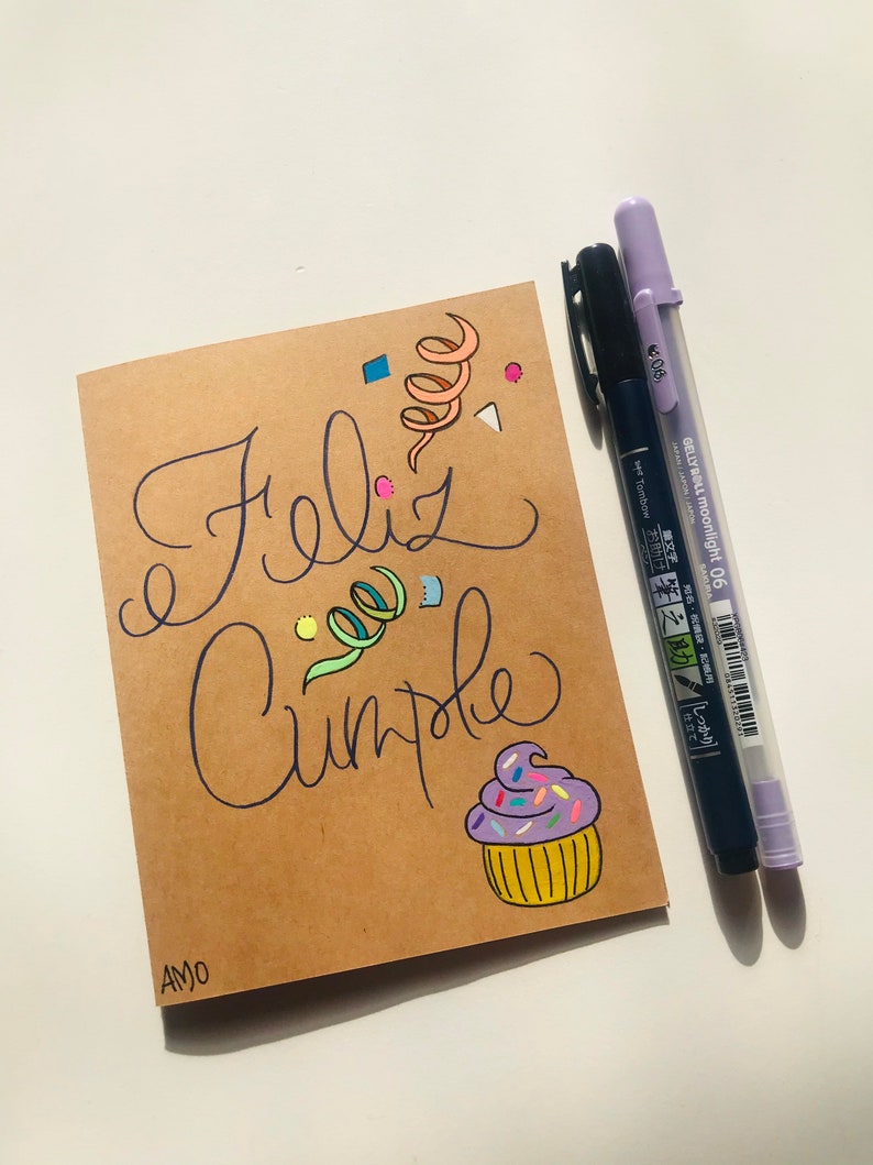 Pu&ograve; includere: Un biglietto marrone con le parole "Feliz Cumple" scritte in inchiostro nero. Il biglietto &egrave; decorato con coriandoli colorati, un vortice di nastro e un cupcake con glassa viola e codette. Il biglietto &egrave; firmato "AMO" nell'angolo in basso a sinistra.
