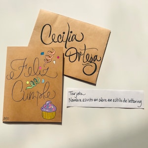 Pu&ograve; includere: Un biglietto marrone con il testo "Feliz Cumple" e un'illustrazione di cupcake colorata. Il biglietto &egrave; decorato con coriandoli e un nome scritto a mano in uno stile di lettering. Il testo "Tarjeta Nombre escrito en sobre en estilo de lettering" &egrave; scritto su un biglietto bianco separato.