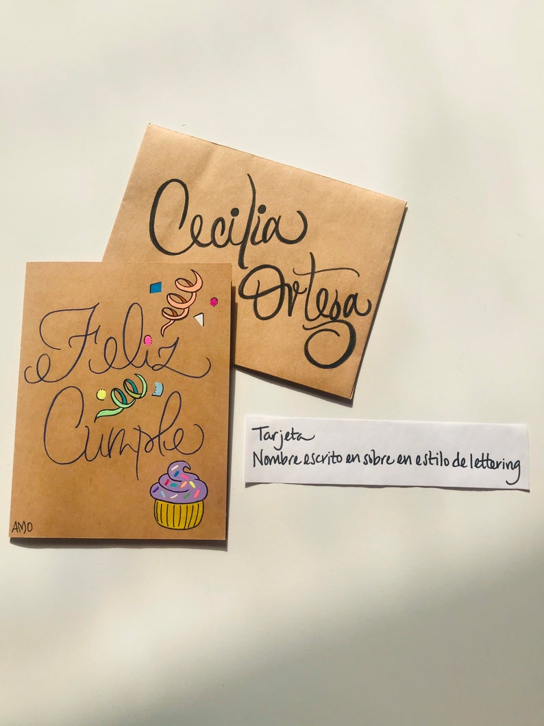 Pu&ograve; includere: Un biglietto marrone con il testo "Feliz Cumple" e un'illustrazione di cupcake colorata. Il biglietto &egrave; decorato con coriandoli e un nome scritto a mano in uno stile di lettering. Il testo "Tarjeta Nombre escrito en sobre en estilo de lettering" &egrave; scritto su un biglietto bianco separato.