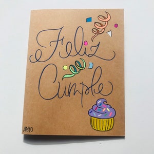 Pu&ograve; includere: Un biglietto marrone con le parole "Feliz Cumple" scritte in inchiostro blu. Il biglietto &egrave; decorato con coriandoli colorati e un cupcake viola con glassa gialla e codette.