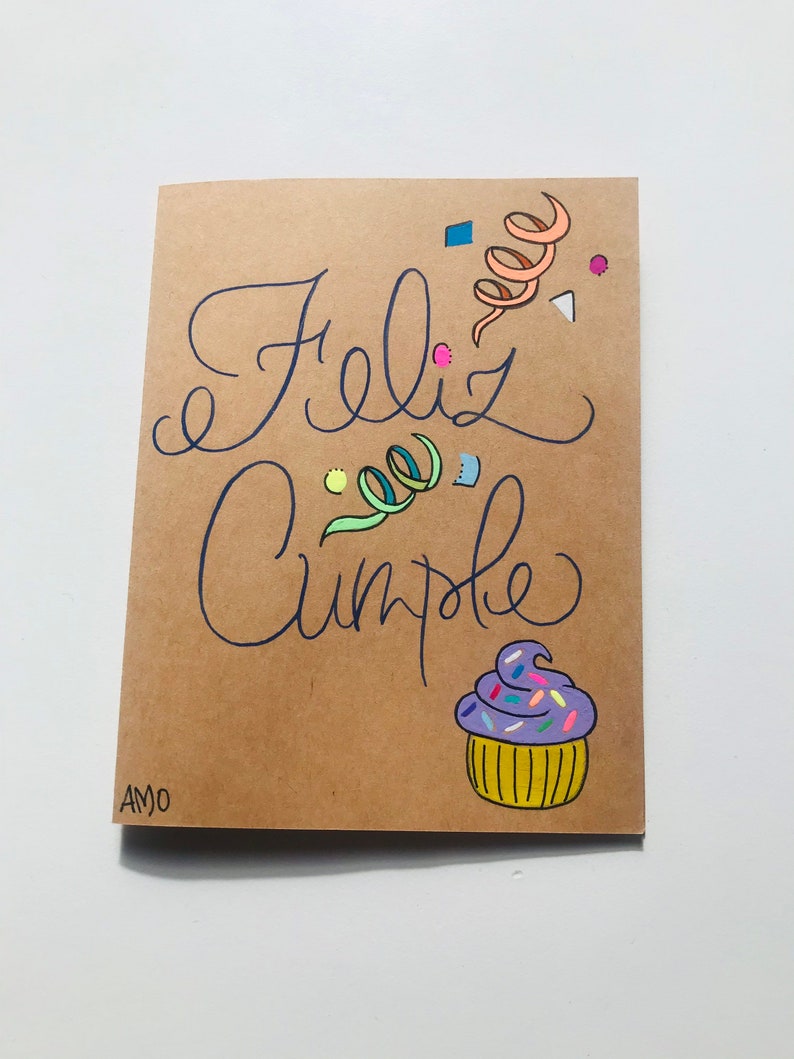 Pu&ograve; includere: Un biglietto marrone con le parole "Feliz Cumple" scritte in inchiostro blu. Il biglietto &egrave; decorato con coriandoli colorati e un cupcake viola con glassa gialla e codette.