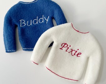 May include: Two felt elf sweaters, one blue with white embroidery that says "Buddy" and one white with red embroidery that says "Pixie".