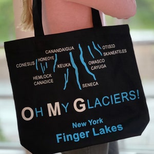 May include: Black canvas tote bag with a blue graphic of the Finger Lakes region of New York State. The graphic shows the shape of the lakes and includes the names of each lake. The text "OH MY GLACIERS!" is printed in white on the bag, along with "New York" and "Finger Lakes" in blue.