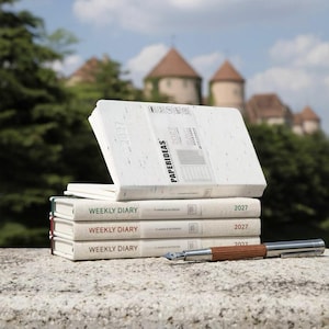 May include: A stack of three off-white weekly diary planners for 2027 with fabric covers and colored spine accents, topped with an open planner featuring a white paper band labeled PAPERIDEAS, accompanied by a silver and brown fountain pen.