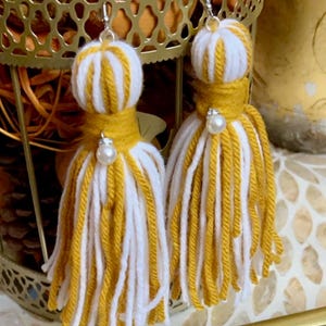 Può includere: Un paio di orecchini a nappa in filato giallo e bianco con accenti di perle. Gli orecchini sono appesi a un supporto in metallo dorato.