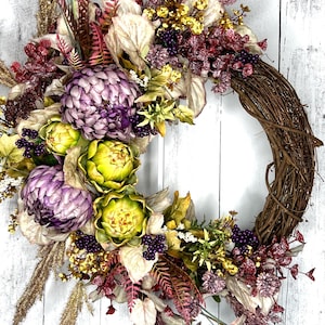 May include: A grapevine wreath decorated with artichoke, berry, and fern accents in shades of purple, green, and gold. The wreath is displayed on a white stand against a whitewashed wood background.
