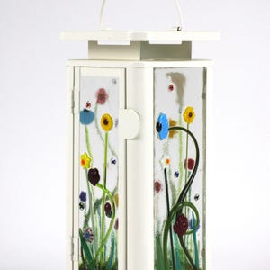 Puede incluir: Una linterna de metal blanco con paneles de vidrio decorados con diseños florales coloridos. La linterna tiene un asa y una pequeña base. Los paneles de vidrio muestran flores en amarillo, azul, rojo y verde, creando una escena de jardín caprichosa. La linterna mide unos 20 cm de alto.