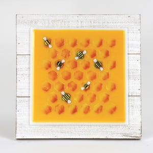 May include: A framed artwork depicting a honeycomb pattern with several small bees. The artwork is set within a white wooden frame. The honeycomb is a bright yellow colour, and the bees have black and yellow stripes.