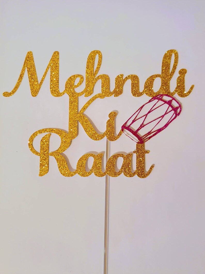 May include: Gold glitter cake topper with the text "Mehndi Ki Raat" and a pink and white dhol drum.