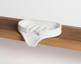 May include: A white ceramic soap dish with a curved design and raised ridges for drainage. The dish is placed on a wooden shelf, showcasing its minimalist aesthetic. The soap dish is designed to hold a bar of soap.